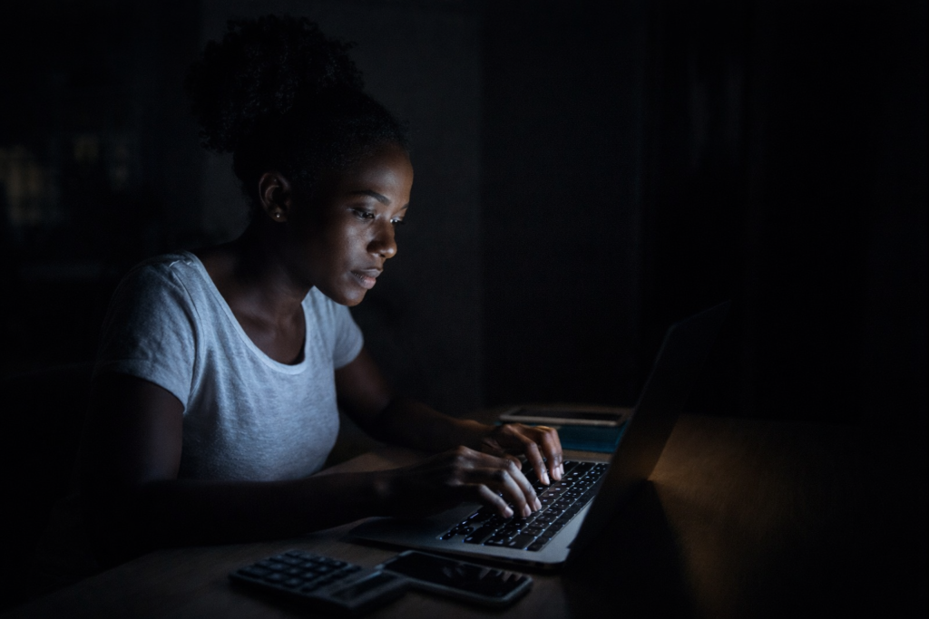 Kenyan woman working at night on laptop with concerned expression, illustrating the high cost of internet access in Africa.”