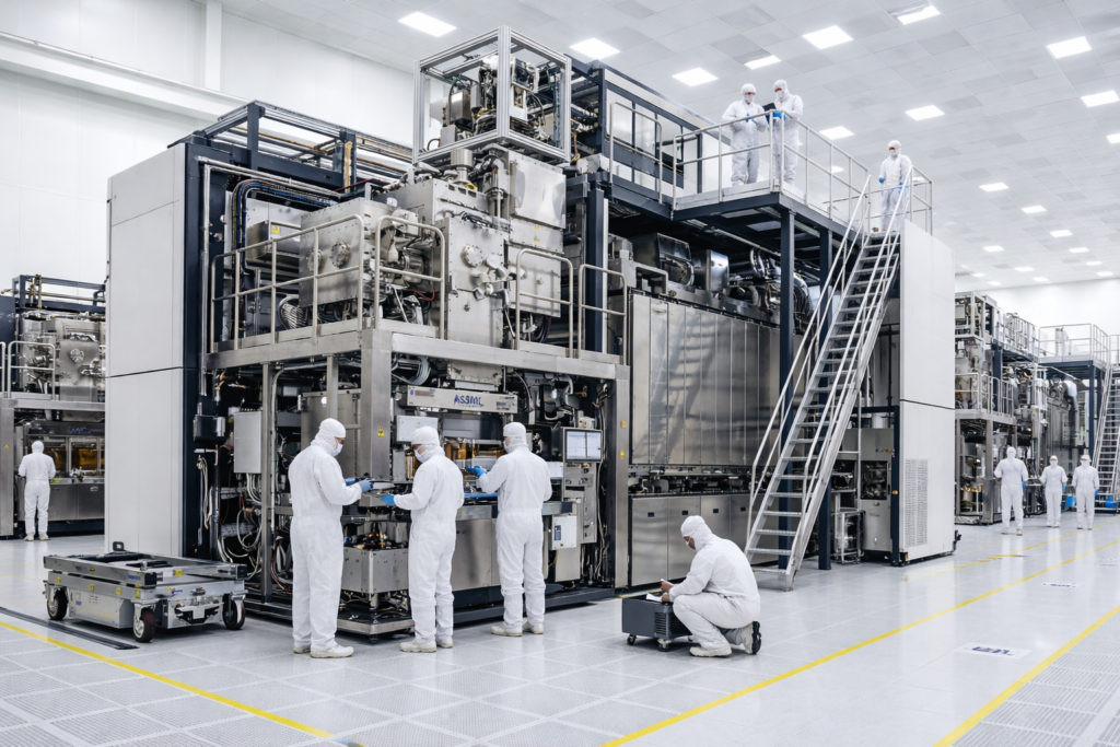 ASML engineers in white clean-room suits work on a Extreme Ultraviolet lithography machine at the company's Veldhoven facility in the Netherlands. The machine towers over the workers, illustrating its 180-metric-ton scale.
